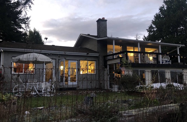 The beach house at dusk, warm light in every window, seen across the garden